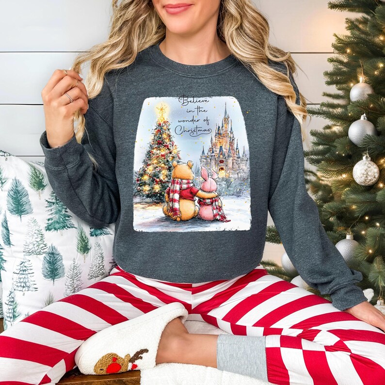 A woman wearing a gray sweatshirt with a Christmas-themed graphic and red and white striped pajama bottoms is sitting on a couch in front of a Christmas tree.