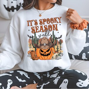 May include: White sweatshirt with the text "It's Spooky Season y'all" and a graphic of a jack-o'-lantern wearing a cowboy hat, boots, and cacti. The sweatshirt is part of a Halloween-themed collection.