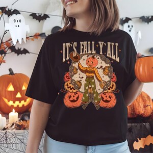 May include: Black t-shirt with the text "IT'S FALL Y'ALL" and a graphic of a scarecrow with a pumpkin head, surrounded by pumpkins and pretzels. The person is holding an orange pumpkin-shaped container.