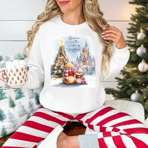 A woman wearing a white sweatshirt with a Christmas-themed graphic and holding a mug of hot chocolate is sitting in front of a Christmas tree.
