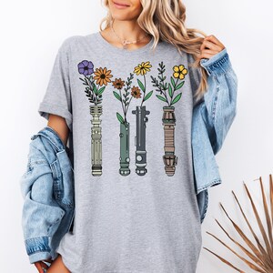 A woman wearing a gray t-shirt with a graphic design of four colorful flowers and plant-like objects, along with a denim jacket.