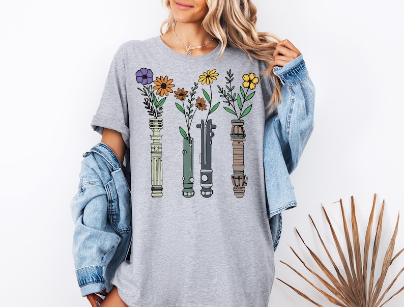 A woman wearing a gray t-shirt with a graphic design of four colorful flowers and plant-like objects, along with a denim jacket.