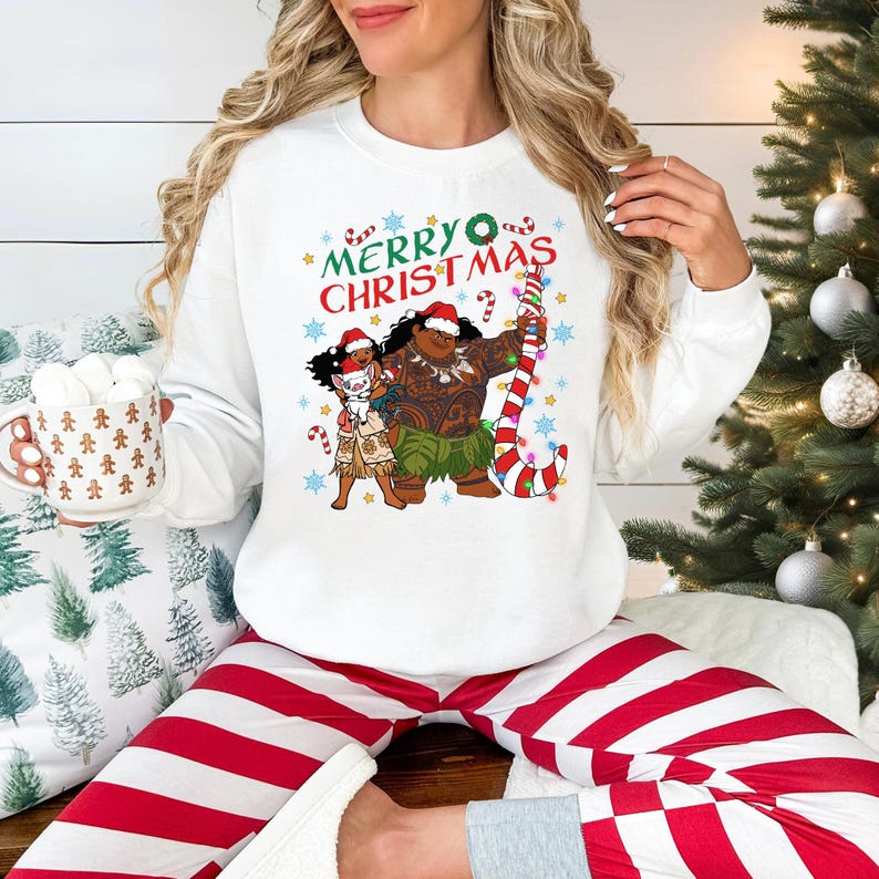 A woman wearing a white sweatshirt with a Christmas-themed graphic and holding a mug is sitting in front of a Christmas tree.