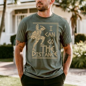 A man wearing a green t-shirt with a graphic design and text printed on it, standing in front of a house with palm trees in the background.