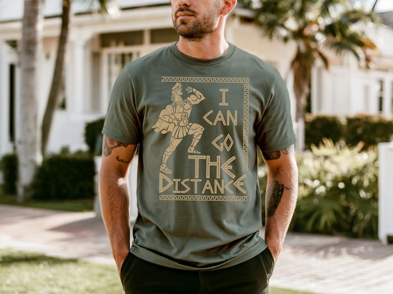 A man wearing a green t-shirt with a graphic design and text printed on it, standing in front of a house with palm trees in the background.