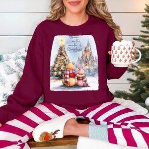 A woman wearing a burgundy sweater with a Christmas-themed graphic and holding a mug is sitting on a couch with striped pajama pants.
