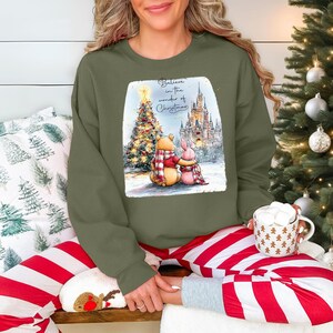 A woman wearing a green sweatshirt with a Christmas-themed graphic and holding a mug of hot chocolate is sitting in front of a Christmas tree.