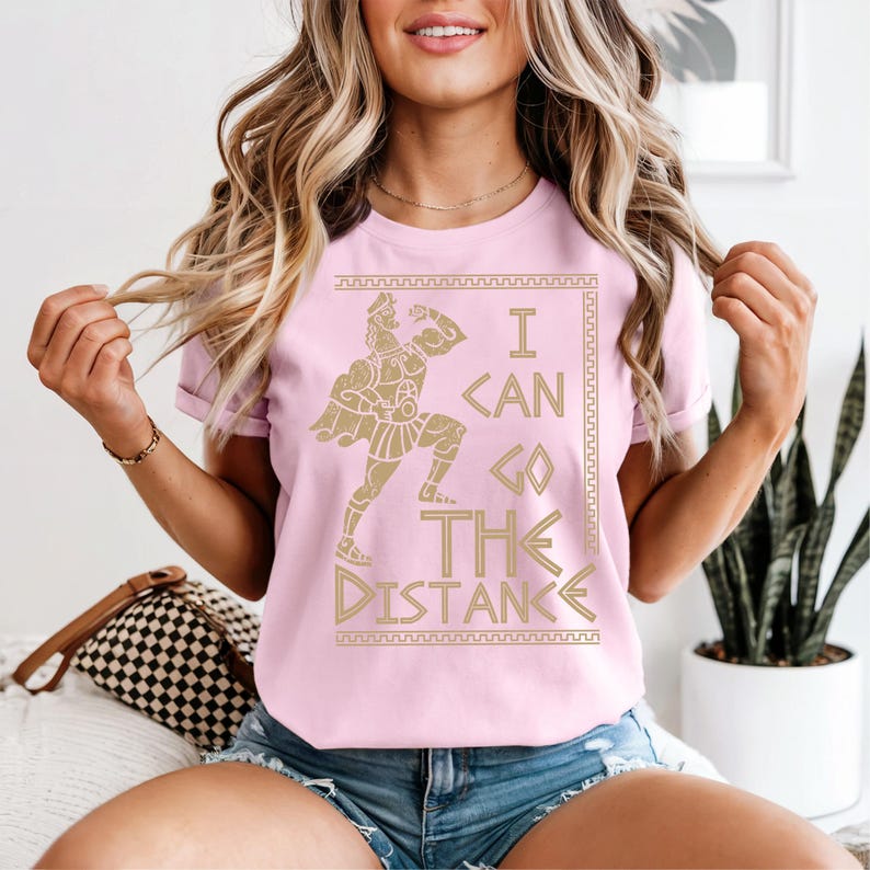 A woman wearing a pink t-shirt with a graphic design and text, sitting in a room with a potted plant in the background.