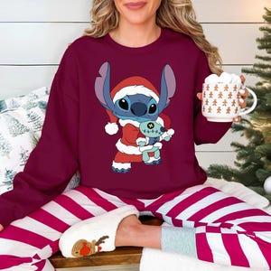 Könnte beinhalten: Burgunderfarbenes Sweatshirt und gestreifte Schlafanzughose mit einer Cartoon-Figur Stitch als Weihnachtsmann, die ein Stofftier hält. Die Person hält einen weißen Becher mit Lebkuchenmännern. Die Hausschuhe haben ein Rentier-Design.