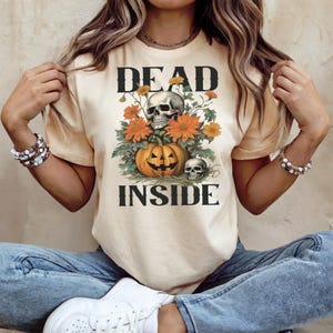 Op de afbeelding: Crèmekleurig T-shirt met de woorden "DEAD INSIDE" in zwarte blokletters. De afbeelding toont een pompoen met een uitgesneden gezicht, twee schedels en oranje bloemen. Het shirt is een casual, Halloween-thema ontwerp.