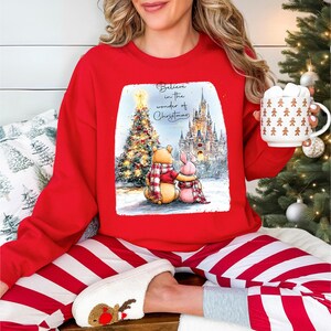 A woman wearing a red sweater with a Christmas-themed illustration is sitting on a bed, holding a mug and surrounded by holiday decorations.