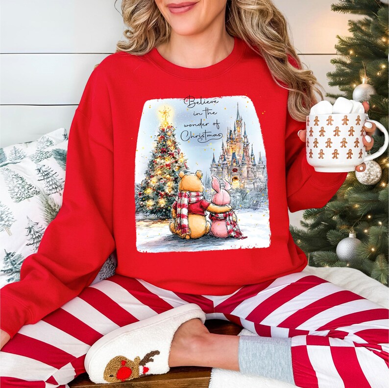 A woman wearing a red sweater with a Christmas-themed illustration is sitting on a bed, holding a mug and surrounded by holiday decorations.