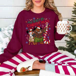 A woman wearing a burgundy sweater with a Christmas-themed graphic and holding a mug is sitting on a couch with striped pajama pants.