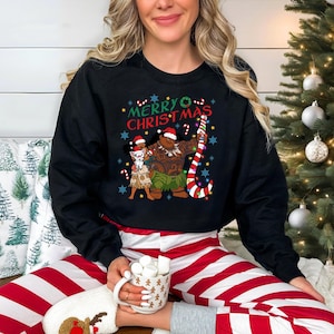 A woman wearing a black sweatshirt with a Christmas-themed graphic and holding a mug of hot chocolate is sitting in front of a Christmas tree.