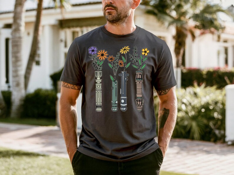 A man wearing a black t-shirt with a graphic design of four vases with flowers on it, standing in front of a house with palm trees.