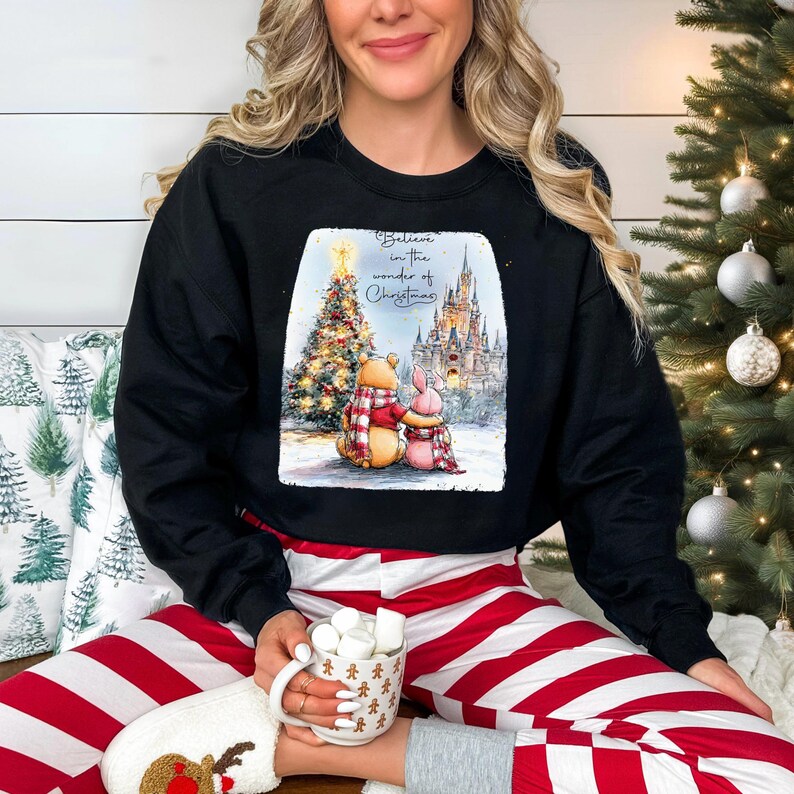 A woman wearing a black sweatshirt with a Christmas-themed graphic is sitting in front of a Christmas tree, holding a mug of hot chocolate.