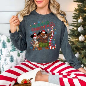 A woman wearing a gray sweatshirt with a Christmas-themed graphic and red and white striped pajama bottoms is sitting in front of a Christmas tree.