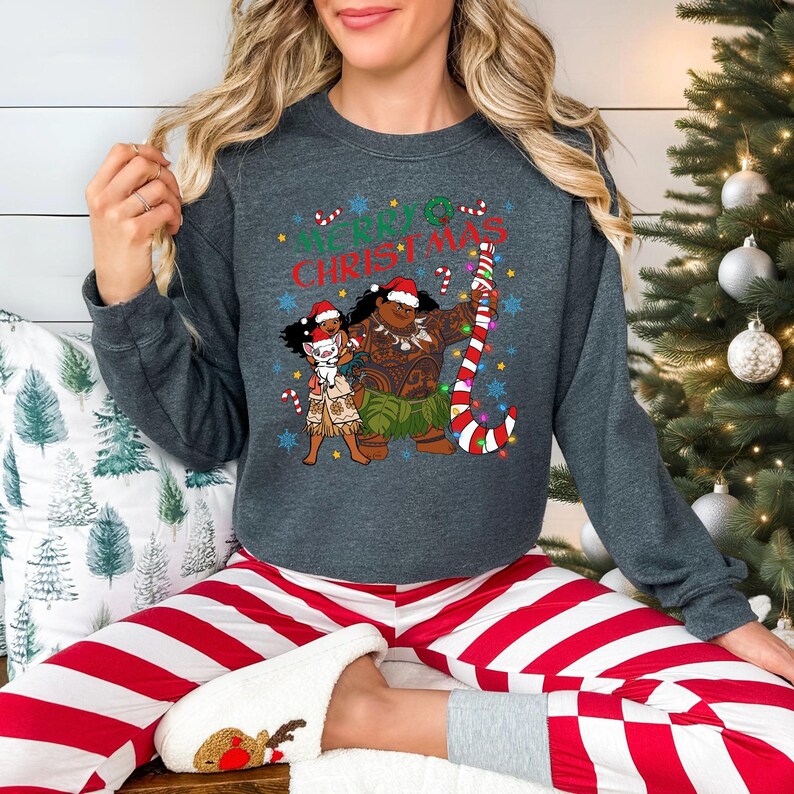 A woman wearing a gray sweatshirt with a Christmas-themed graphic and red and white striped pajama bottoms is sitting in front of a Christmas tree.