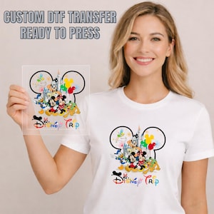 May include: White t-shirt with a colourful Disney Trip graphic. The design features Mickey Mouse, Minnie Mouse, Donald Duck, Goofy, and other characters in front of a castle. The image also shows a clear DTF transfer with the text "CUSTOM DTF TRANSFER READY TO PRESS".
