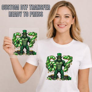 May include: A white t-shirt featuring a green gorilla holding the numbers "67" with a St. Patrick's Day theme. The numbers are green and white striped, and the gorilla is wearing a green hat. The text "CUSTOM DTF TRANSFER READY TO PRESS" is at the top.