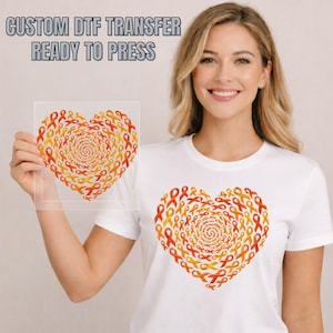 May include: A white t-shirt featuring a heart-shaped graphic composed of orange and yellow awareness ribbons arranged in a spiral pattern. A matching square DTF transfer sheet with the same heart design is held up, with text reading CUSTOM DTF TRANSFER READY TO PRESS.