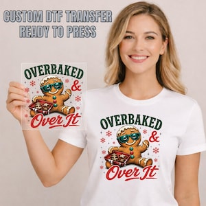 May include: A white t-shirt featuring a graphic of a gingerbread person wearing teal sunglasses, holding a baking tray of cookies, with the text OVERBAKED & OVER IT. The design includes red snowflakes and green accents, printed as a DTF transfer.