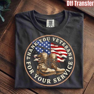 May include: Dark grey t-shirt with a circular graphic. The graphic features an American flag, combat boots, and the text "THANK YOU VETERANS FOR YOUR SERVICE" in a circular design. The shirt has a tag that reads "COMFORT COLORS".