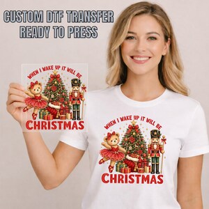 May include: A white t-shirt featuring a graphic print of a teddy bear in a red tutu, a nutcracker, a decorated Christmas tree, and wrapped gifts. The text reads, "WHEN I WAKE UP IT WILL BE CHRISTMAS." A matching square DTF transfer sheet is also shown.