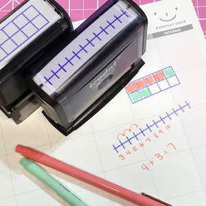 May include: Two stamps, one with a grid and one with a ruler, are on a white gridded paper with two colored pens. The paper has a handwritten math problem, "4 + 3 = 7", and a number line with numbers 3 through 10. The paper also has a grid with red and green squares. The paper has a header that says "EVERYDAY SMILE SATURDAY".