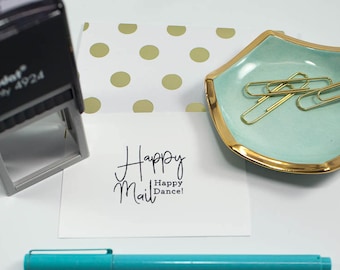 May include: A white card with the text "Happy Mail Happy Dance!" written in black ink. A turquoise blue pen, a gold and teal dish with gold paper clips, and a black stamp are also in the image.