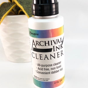 May include: A white bottle of archival ink cleaner with a black cap and a rainbow label. The bottle is labeled "Archival Ink Cleaner" and lists the product features: "All-purpose cleaner", "Acid free, non toxic", and "Convenient dabber top". The bottle contains 2 fluid ounces (59 ml).