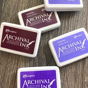Peut inclure: Cinq tampons d'encre dans des &eacute;tuis en plastique blanc avec la marque "Ranger" et le texte "Archival Ink" en noir. Les tampons d'encre sont de couleur violet, prune, chardon, violet et violet majestueux.