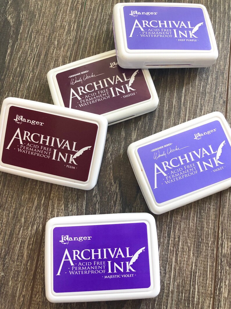 Peut inclure: Cinq tampons d'encre dans des &eacute;tuis en plastique blanc avec la marque "Ranger" et le texte "Archival Ink" en noir. Les tampons d'encre sont de couleur violet, prune, chardon, violet et violet majestueux.