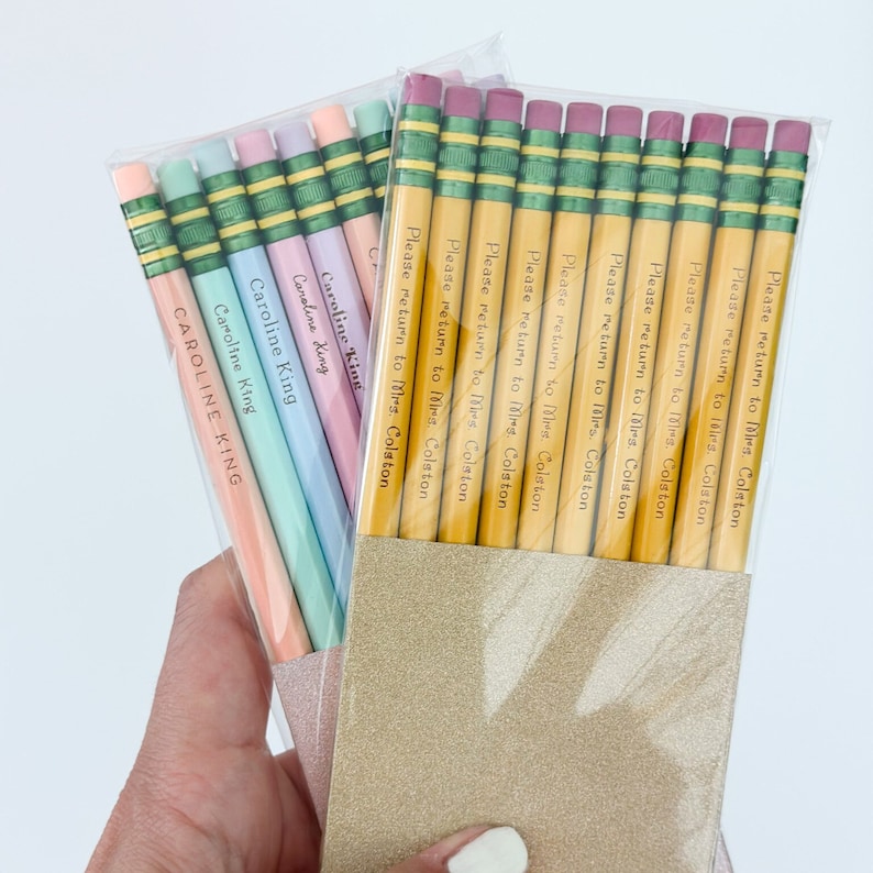 May include: Two pencil sets in clear packaging. One set features pastel pencils with "Caroline King" printed on them. The other set has yellow pencils with "Please return to Mrs. Colston" printed on them. Both sets are in a gold glittery holder.