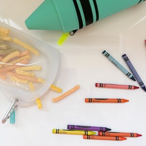 May include: A teal crayon case with black stripes and a zipper. The case is open and crayons are scattered on a white surface. The crayons are in various colors, including red, orange, yellow, green, blue, purple, and pink.