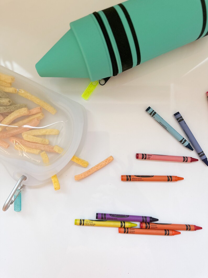 May include: A teal crayon case with black stripes and a zipper. The case is open and crayons are scattered on a white surface. The crayons are in various colors, including red, orange, yellow, green, blue, purple, and pink.