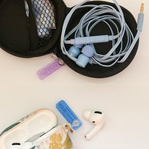 May include: A floral patterned case for wireless earbuds with a blue and white personalized name tag. The case is open and the earbuds are visible. A separate black case with a blue cord and earbuds is also shown.