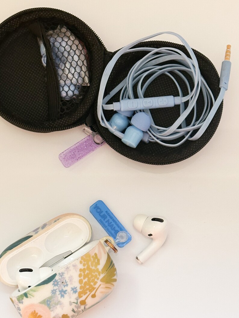 May include: A floral patterned case for wireless earbuds with a blue and white personalized name tag. The case is open and the earbuds are visible. A separate black case with a blue cord and earbuds is also shown.