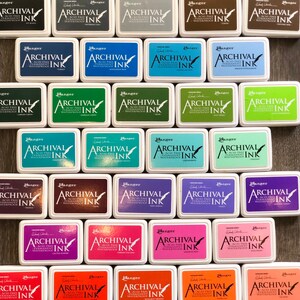 Peut inclure: Une collection de 36 tampons encreurs de diff&eacute;rentes couleurs. Chaque tampon porte l'inscription "Ranger Archival Ink" et est &eacute;tiquet&eacute; avec le nom de la couleur. Les tampons encreurs sont dispos&eacute;s en rang&eacute;es et en colonnes sur une surface en bois.