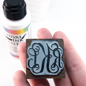 May include: A wooden stamp with a black ink design of the letters "DA" in a decorative script font. The stamp is being held in a hand. A bottle of archival ink is visible in the background.
