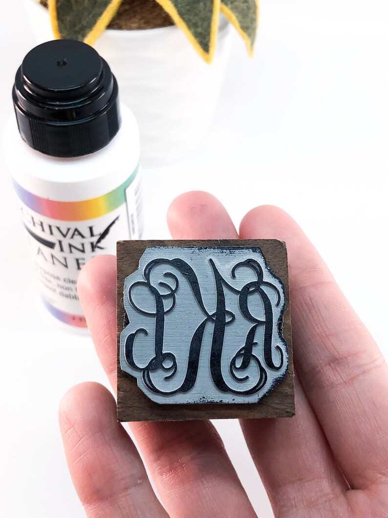 May include: A wooden stamp with a black ink design of the letters "DA" in a decorative script font. The stamp is being held in a hand. A bottle of archival ink is visible in the background.
