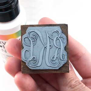 May include: A close-up of a wooden stamp with a gray monogram design. The monogram is a stylized "MA" with a decorative flourish.