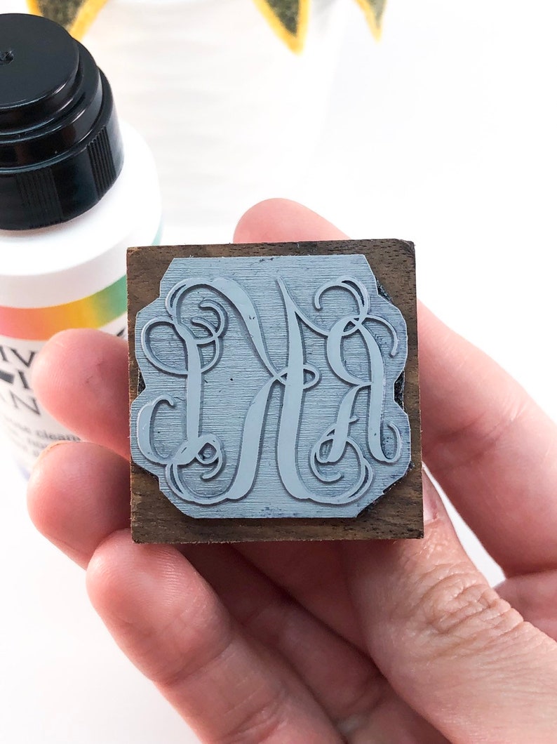 May include: A close-up of a wooden stamp with a gray monogram design. The monogram is a stylized "MA" with a decorative flourish.