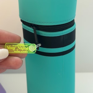 May include: A turquoise blue crayon-shaped pencil case with black stripes and a zipper closure. A yellow plastic tag attached to the zipper reads "PLEASE RETURN TO Your Name PHONE NUMBER".