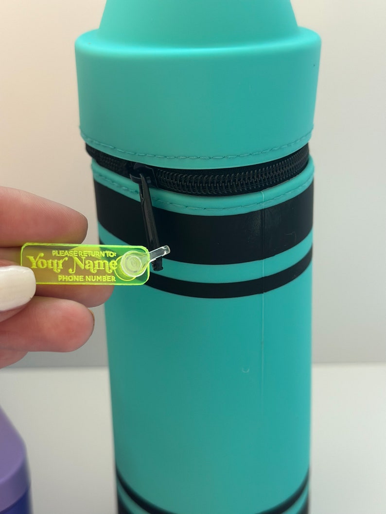 May include: A turquoise blue crayon-shaped pencil case with black stripes and a zipper closure. A yellow plastic tag attached to the zipper reads "PLEASE RETURN TO Your Name PHONE NUMBER".