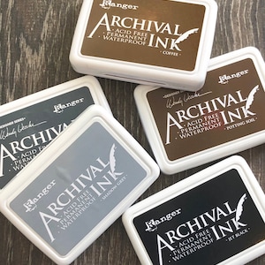 Peut inclure: Quatre tampons d'encre avec des &eacute;tiquettes blanches et du texte noir. Les &eacute;tiquettes indiquent "Ranger Archival Ink" et listent la couleur de chaque tampon d'encre : "Coffee", "Shadow Grey", "Potting Soil", et "Jet Black".