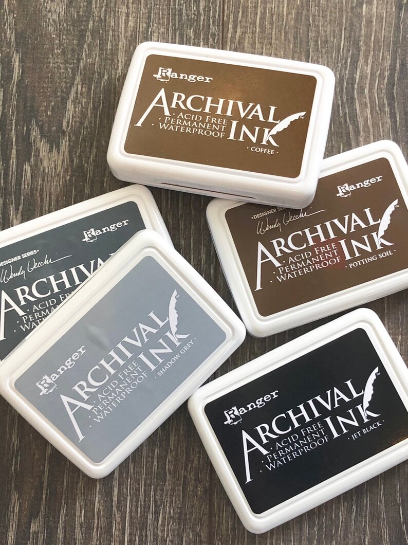 Peut inclure: Quatre tampons d'encre avec des &eacute;tiquettes blanches et du texte noir. Les &eacute;tiquettes indiquent "Ranger Archival Ink" et listent la couleur de chaque tampon d'encre : "Coffee", "Shadow Grey", "Potting Soil", et "Jet Black".