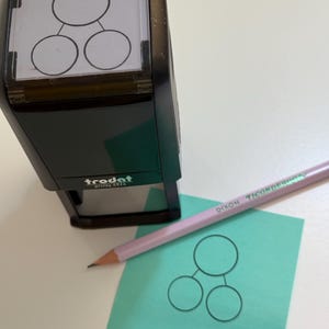 May include: A black self-inking stamp with a clear window showing a diagram of three circles connected by lines. The stamp is labeled "trodat printy 4924". A pencil is lying next to a green sticky note with a similar diagram drawn on it.