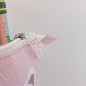 May include: A pink canvas pencil case with a white cat face design. The zipper pull has a clear plastic tag with the text "Little Fox" printed on it.