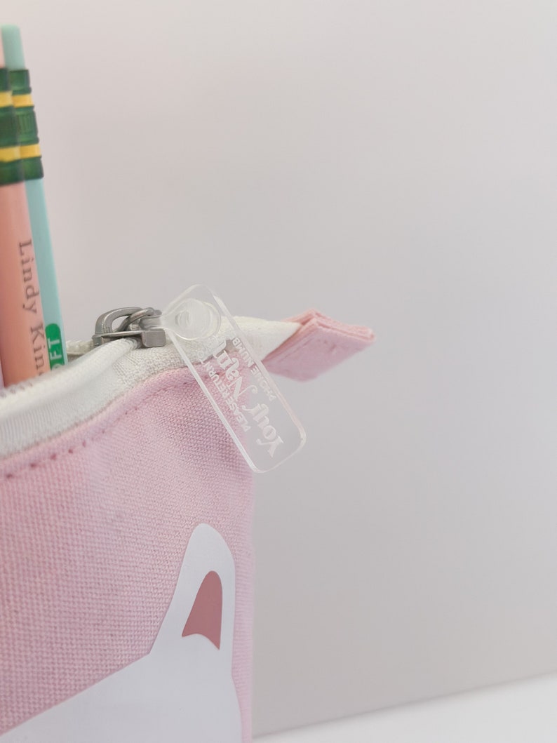 May include: A pink canvas pencil case with a white cat face design. The zipper pull has a clear plastic tag with the text "Little Fox" printed on it.